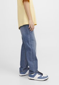 Ljusgul bomulls t-shirt med avslappnad passform. Blå denimjeans med rak skärning. Blå och vita sneakers med svarta skosnören och texturerade sulor.