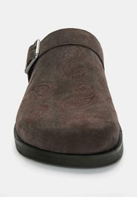 Brązowy zamszowy klapek z wytłaczanym wzorem paisley; posiada metalową klamrę oraz czarną gumową podeszwę. Kształt jest okrągłoznosy i o niskim profilu.