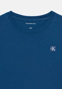 Blue cotton t-shirt with a classic crew neck. Features a small CK logo in a square on the left chest area. Smooth texture, short sleeves.