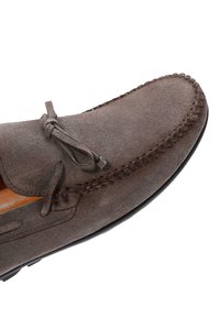 Zapato mocasín de ante marrón con un detalle cosido a lo largo del borde, que presenta un lazo y una suela de goma negra lisa.