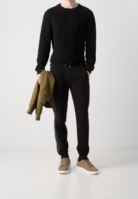 Suéter negro de canalé, pantalones deportivos negros con cordón, sosteniendo una sudadera con capucha verde oliva y usando zapatillas de ante color marrón claro. Diseño simple y casual.
