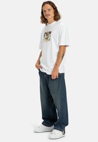 Camiseta de algodón blanca con un gráfico floral, combinada con jeans de mezclilla azul holgados y zapatillas blancas. Cuenta con un corte relajado y mangas cortas.