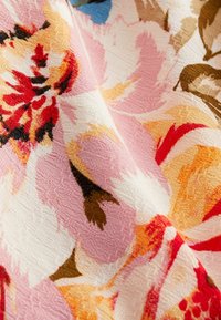 Tissu floral présentant de grandes fleurs roses, orange et rouges sur un fond crème. Surface texturée avec un motif de tissage visible.