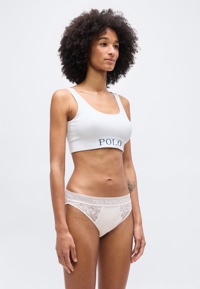 Γυναίκα με σγουρά μαλλιά που φοράει ένα λευκό αθλητικό σουτιέν με την επιγραφή "POLO" και ανοιχτό ροζ εσώρουχο με δαντέλα, στέκεται μπροστά σε μια απλή φόντο.