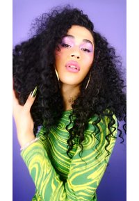 Model with long, curly black hair wearing a green and yellow patterned long-sleeve top against a purple background. Gold hoop earrings.