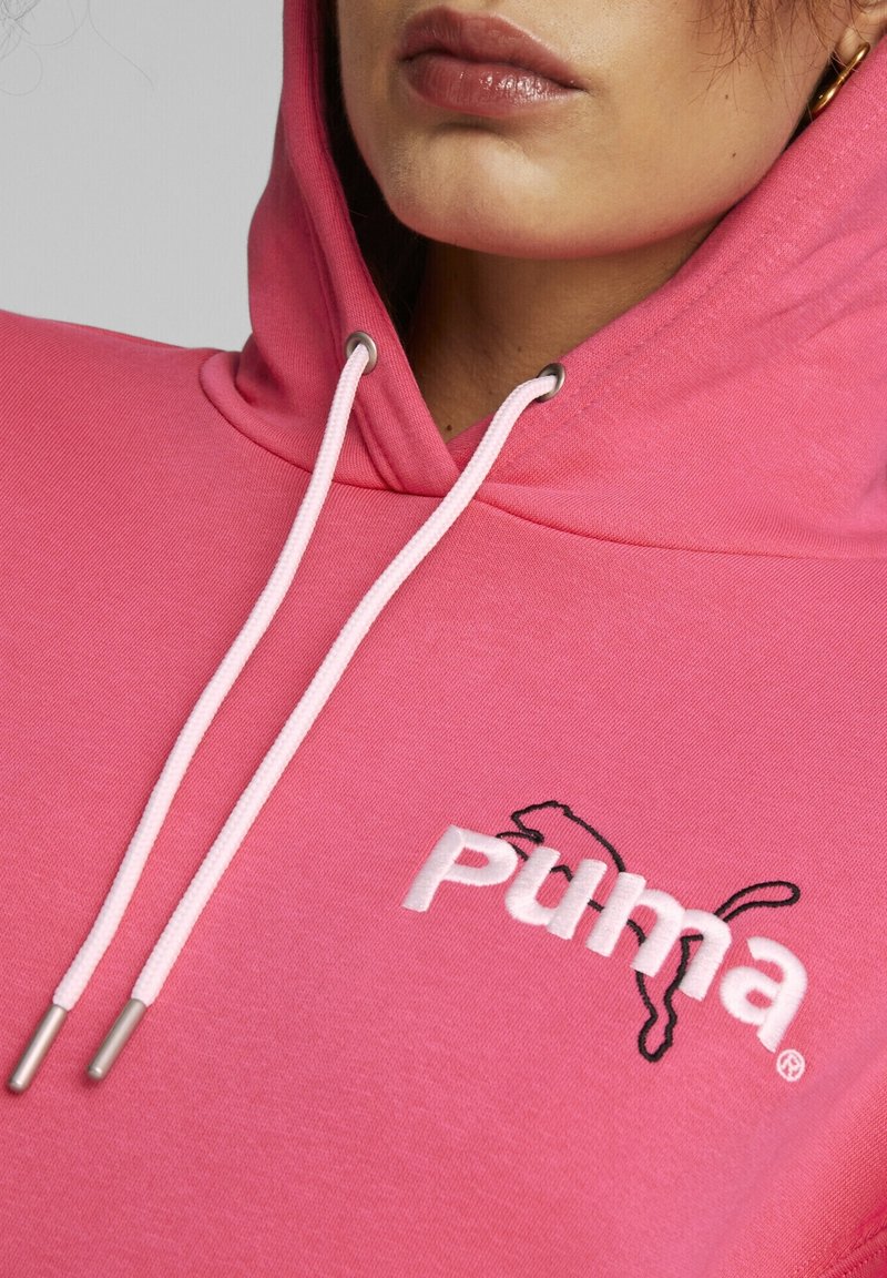 Sudadera rosa con un bolsillo canguro frontal, cordones blancos y logo bordado en blanco y negro de Puma en el pecho. Textura del tejido suave.