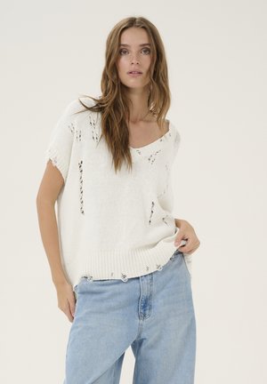 Pull en tricot blanc avec un tissage ouvert et des détails déchirés, manches courtes et ourlet côtelé. Associé à un jean bleu clair à jambe droite.