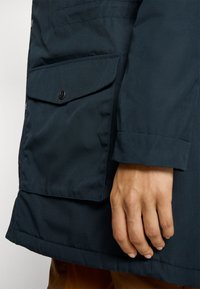 Marineblauwe jas met een voorzak met drukknoopsluiting, zichtbare stiksels en een gestructureerde stof. Gefronte mouwen.