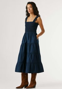 Nő pánt nélküli tengerészkék réteges ruhában, fodros pántokkal és barna velúr cowboy csizmában, egyik kezét a zsebében tartva áll.