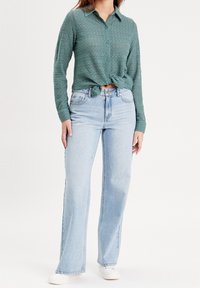 Chemise verte à manches longues texturée, nouée à la taille, associée à un jean large en denim bleu clair et des baskets blanches.