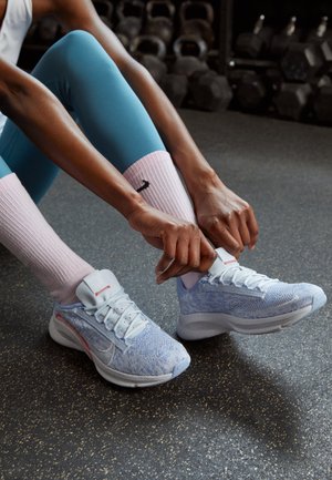 Personne en leggings bleus et chaussettes roses nouant des chaussures de course bleu clair sur le sol de la salle de sport avec des haltères en arrière-plan.