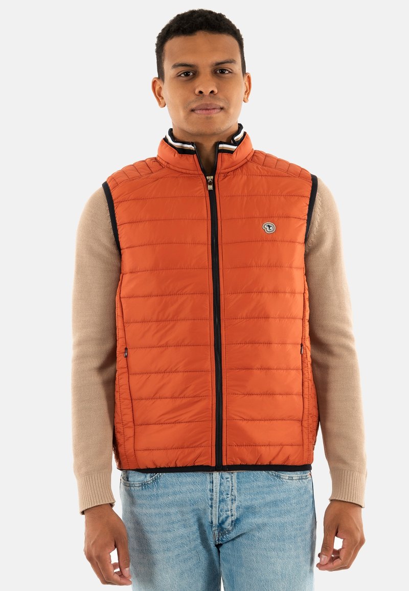 Gilet matelassé orange avec fermeture éclair sur le devant, col montant et bordures bleu marine. Porté par-dessus un pull beige et associé à un jean bleu clair.