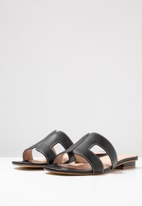 Black synthetic slide sandals with open toe, featuring a cut-out design and contrasting white stitching on the upper. Flat sole.