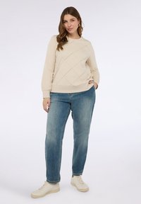 Béžová prošívaná mikina s dlouhými rukávy, kulatým výstřihem a žebrovaným spodním lemem, v kombinaci se světle modrými rovnými džínami a bílými teniskami.