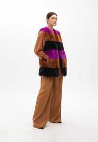 Faux fur vest with bold horizontal stripes in magenta, black, and brown, featuring a wide collar and pockets. Paired with brown wide-leg trousers.