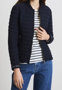 Navy quilted bomber jacket with a zip closure, ribbed collar, and contrasting cuffs. Worn over a striped shirt and paired with blue jeans.