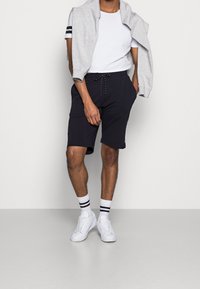 Černé sportovní šortky se šňůrkou, v kombinaci s bílým tričkem a šedou mikinou přehozenou přes ramena. Bílé tenisky a pruhované ponožky.