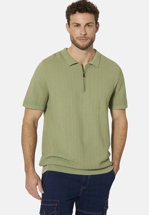 Junger Mann trägt ein hellgrünes kurzärmeliges Strickpoloshirt mit einem halben Reißverschluss am Kragen und dunkelblaue Cargohosen.