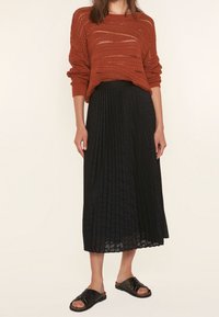 Rostfärgad stickad tröja med lös passform och texturerat mönster, kombinerad med en svart plisserad midiklänning och svarta platta sandaler.