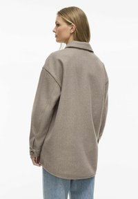 Overdimensioneret brun uld skjortejakke med krave, nedfaldne skuldre, knaplukninger ved håndleddene og en buet kant, båret med lyseblå jeans.