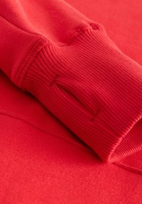 Czerwona bluza z teksturowanym ściągaczem i miękką, polarową wyściółką, z gładkim zewnętrznym wykończeniem. Widoczne przeszycia wzdłuż szwów.