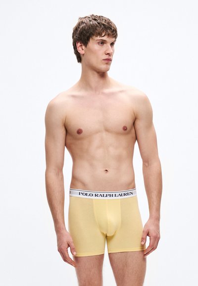 Svetlo rumene bokserice z belim pasom, na katerem je črn napis "POLO RALPH LAUREN". Gladek material in oprijeta zasnova.