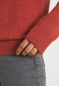 Käsi osaliselt punase kudumise väljanägemisega jakikrae all, puhkamas hallide teksade taskuõmbluse lähedal.