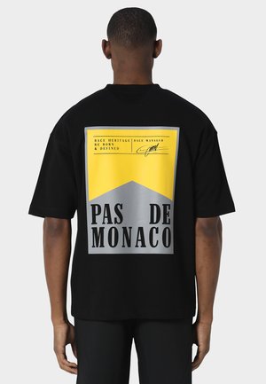 Uomo che indossa una t-shirt nera oversize con una grande grafica rettangolare gialla e grigia con la scritta "Pas De Monaco" sulla schiena su sfondo semplice.