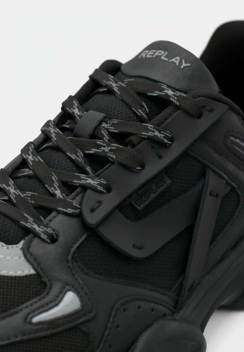 Basket noire et grise Replay avec des lacets à motifs, tige en mesh et synthétique, et logo de marque visible sur la languette et le panneau latéral.