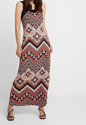 Mujer con un vestido largo sin mangas con patrones geométricos y florales en negro, rojo, blanco y marrón, combinado con sandalias de tacón alto negras con tiras.