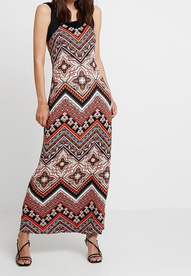 Femme portant une robe maxi sans manches avec des motifs géométriques et floraux noirs, rouges, blancs et marron, associée à des sandales noires à talons hauts et à brides.