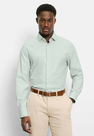 Mann trägt ein hellgrünes Hemd mit Knöpfen, das in beige Hose mit braunem Gürtel gesteckt ist, steht mit einer Hand in der Tasche.