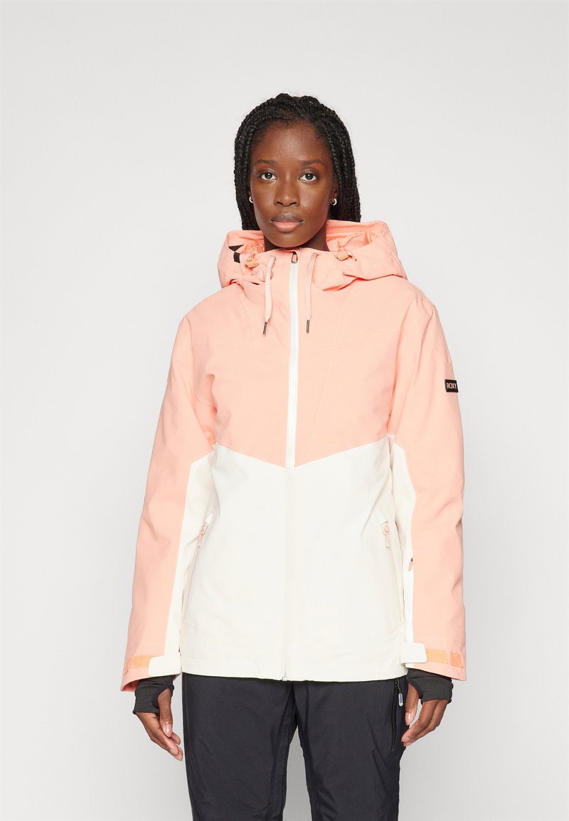 Giacca impermeabile in pesca e bianco con cappuccio, due tasche laterali con zip e cordini regolabili. Presenta un design geometrico a blocchi di colore.