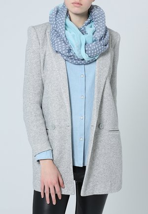 Personne portant un blazer gris clair, une chemise boutonnée bleu clair, un pantalon noir et une écharpe bleue à motifs d'étoiles blanches.