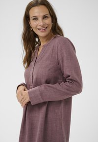 Smeška ženska z dolgimi rjavimi lasmi nosi mauve obleko z dolgimi rokavi in brez ovratnika, stoječa pred enostavno svetlo ozadje.