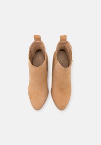 Botas de tornozelo em camurça bege com um design elegante, apresentando um painel elástico lateral, uma alça para puxar e uma sola de borracha texturizada para melhor aderência.