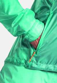 Grüne, leichte Jacke mit glatter Textur, ausgestattet mit einer Reißverschlusstasche und einem leuchtend orangefarbenen Reißverschlusszieher. Elastische Bündchen und Saum.