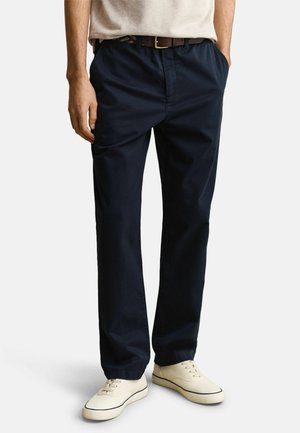 Mann trägt dunkelblaue gerade geschnittene Hose, weiße Turnschuhe, braunen Gürtel und hellbeiges Hemd, Hände in den Taschen, steht vor weißem Hintergrund.