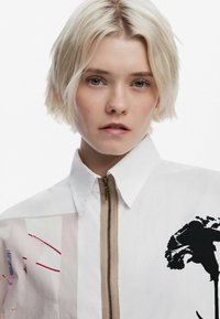 Chemise blanche avec fermeture éclair à l'avant, arborant un grand design graphique noir et des motifs colorés abstraits dans des teintes de rose et de rouge d'un côté.