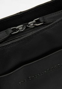 Mustan nahkalaukun, jossa on ylhäältä avattava vetoketju ja tekstuuri, etupuolella on hienovaraisesti kohokuvioitu logo ja metalliset yksityiskohdat.