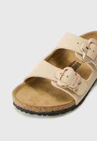 Beige sandal med två justerbara remmar och metalliska spännen i form av blommor. Korkfotbädd och texturerad gummisul för bra grepp.