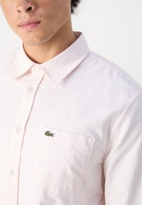 Camicia button-down rosa chiaro con sottili strisce bianche. Presenta un colletto classico, maniche lunghe e tasca con logo ricamato verde.