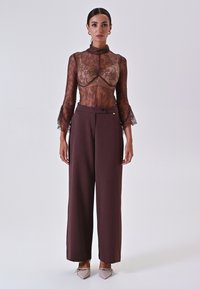 Top in pizzo con colletto alto e maniche a campana in trasparente marrone, sovrapposto a un pantalone a vita alta ampio e solido di un marrone scuro, con dettaglio a pulsante laterale.