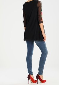 Schwarze Tunika mit durchsichtigen langen Ärmeln und floralen Stickereien, kombiniert mit blauen Skinny Jeans und roten High Heels.
