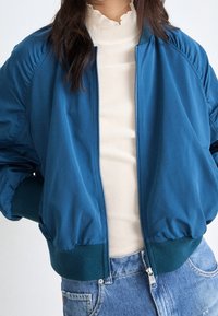 Teal bomberjacka med dragkedja fram, ribbade ärmslut och nederkant. Bärs över en krämfärgad skjorta, kombinerad med denimshorts. Slät textur och avslappnad passform.