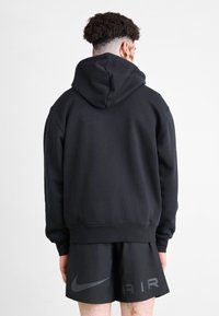 Man with curly hair wearing a black hooded sweatshirt and black Nike Air shorts, standing facing away against a plain background.