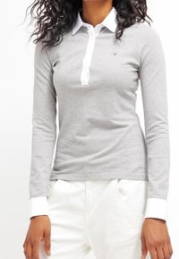 Camisa polo de manga larga gris claro, hecha de una tela suave, con un cuello y puños blancos, y un pequeño logotipo en el área del pecho izquierdo.