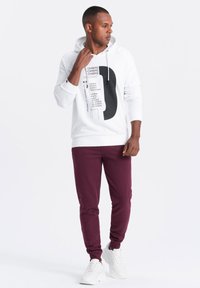 Weißer Hoodie mit schwarzem Grafikdruck, Kängurutasche und Kapuze mit Kordelzug, kombiniert mit bordeauxroten Jogginghosen und weißen Sneakers.