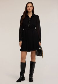 Schwarzes, plissiertes Kleid mit langen Ärmeln, einem kragenförmigen Ausschnitt und einem subtilen Knopfdetail. Kombiniert mit knielangen schwarzen Stiefeln und einer kleinen Handtasche.