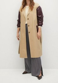 Cappotto lungo beige con maniche in pelle color bordeaux, chiusura doppiopetto con bottoni, tasche laterali e tessuto a contrasto. Indossato sopra pantaloni stampati.